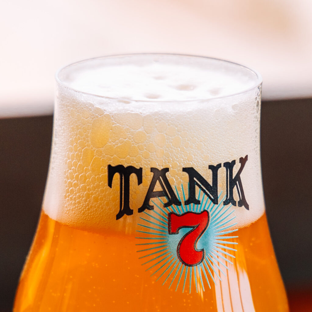 A close-up shot of a beer glass filled with golden beer, topped with a frothy white head. The glass is branded with the word "TANK" in bold letters and a red number "7" with a blue starburst design behind it.
