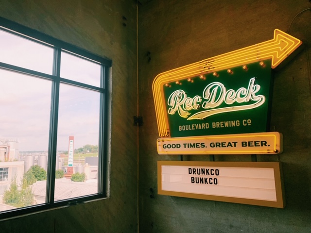 A neon sign reading "Rec Deck Boulevard Brewing Co" with "Good Times. Great Beer." underneath, and a message board with "Drunkco Bunkco." A window shows an industrial view outside.