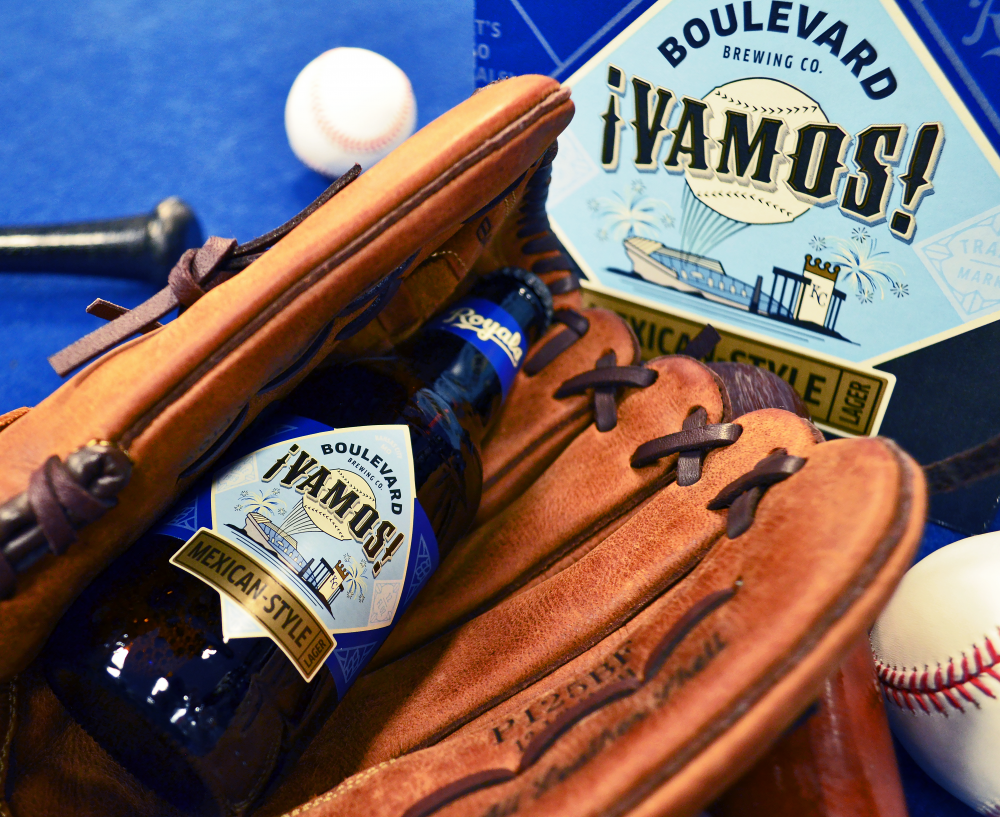 A bottle of Boulevard '¡Vamos!' Mexican-style beer rests inside a brown baseball glove. Another glove, a baseball bat, and two baseballs are nearby. The beer label shows a ship and the brand name, hinting at a baseball theme.