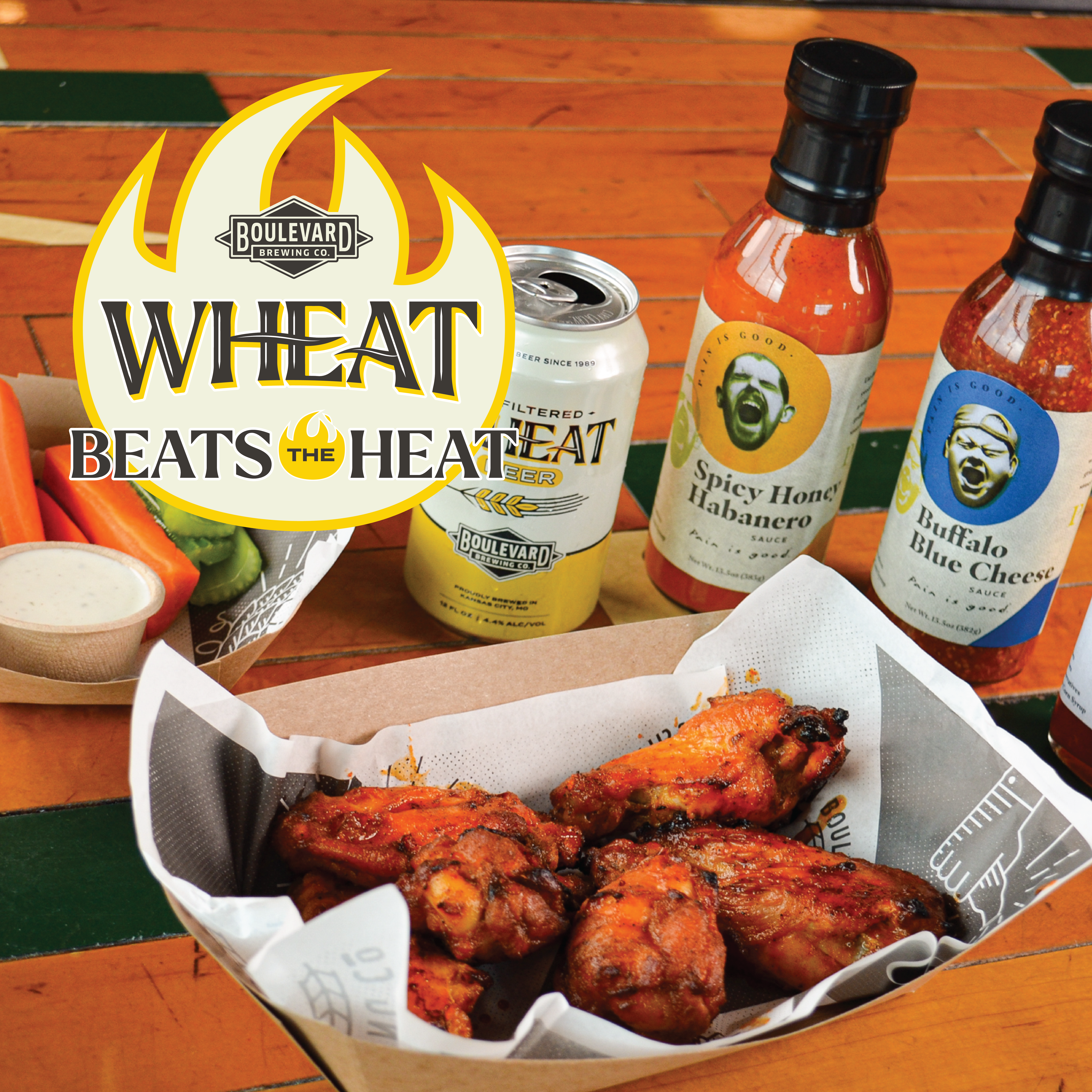 A close-up of a table with buffalo chicken wings in a basket, accompanied by dipping sauce, carrot and celery sticks, and hot sauce bottles. A can of Boulevard Wheat beer is also present, along with the slogan "Wheat Beats the Heat" and a logo featuring flames.