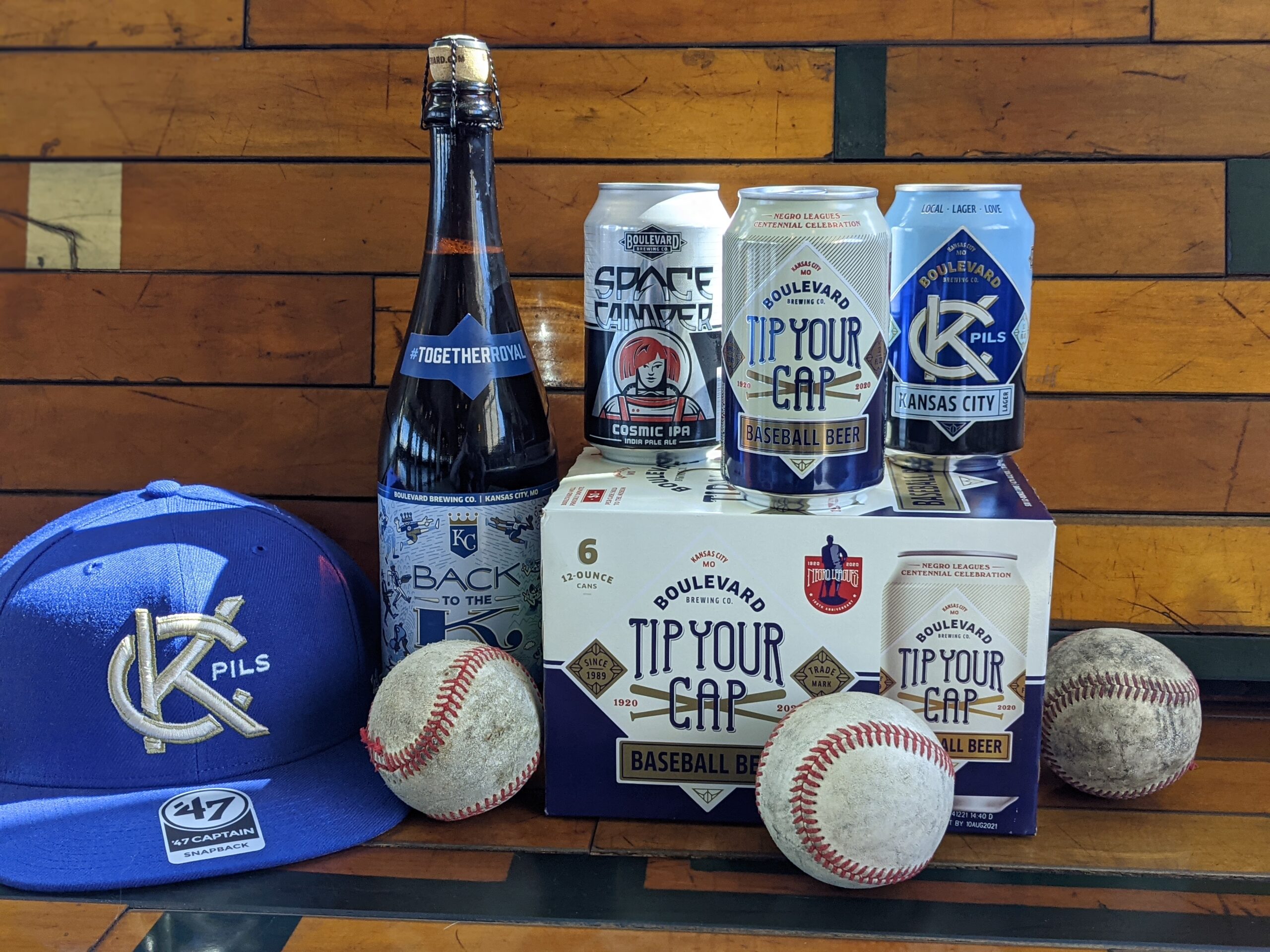 A display of various Boulevard Brewing Company beer products and baseball paraphernalia. The beers include a bottle and cans labeled "KC Pils" and "Tip Your Cap." A blue baseball cap with a letter "KC" logo and three baseballs surround the items.