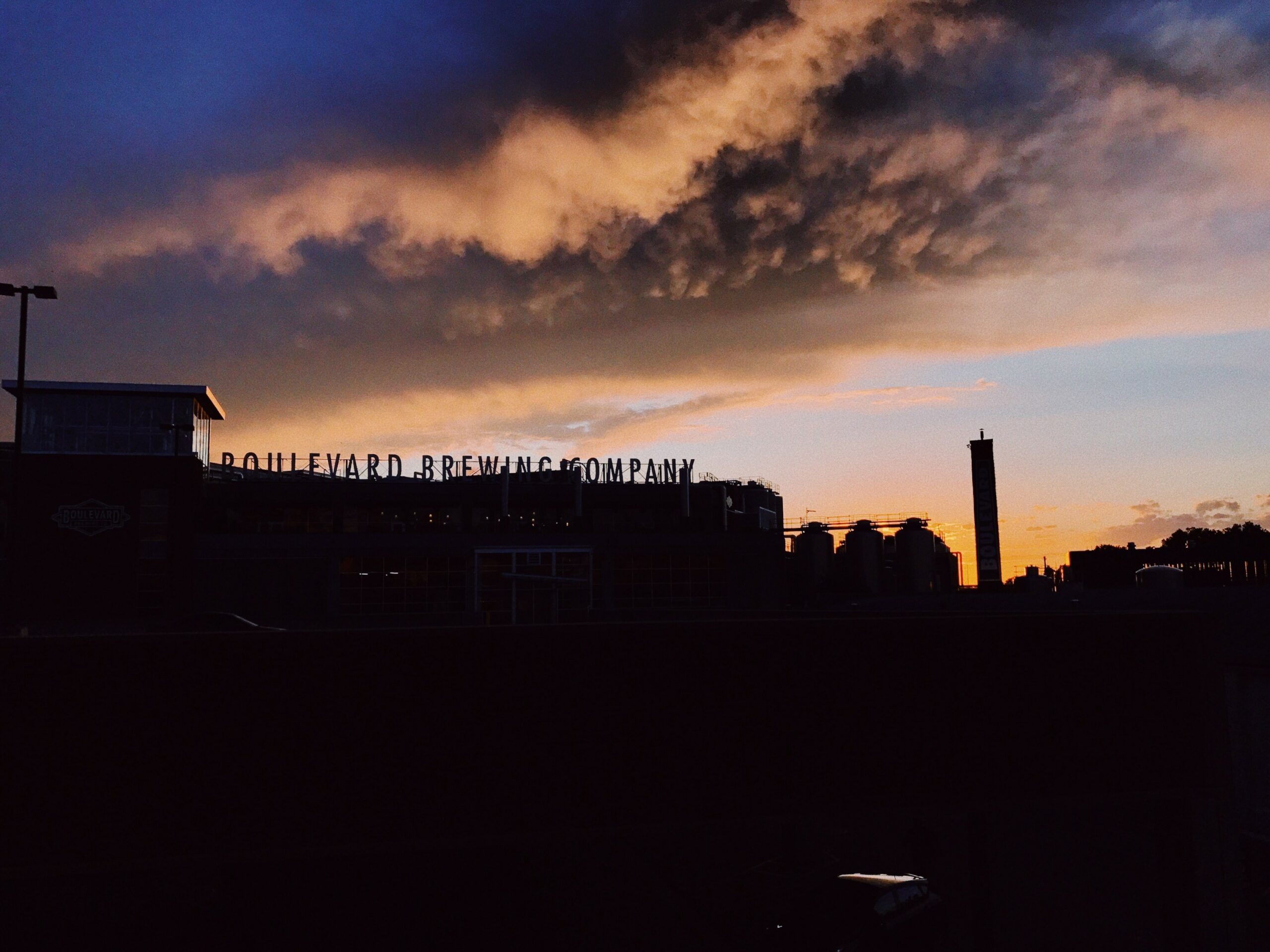 Sunset view over Boulevard Brewing Company, with dramatic clouds and an orange sky creating a silhouette of the brewery buildings and a tall chimney in the background.