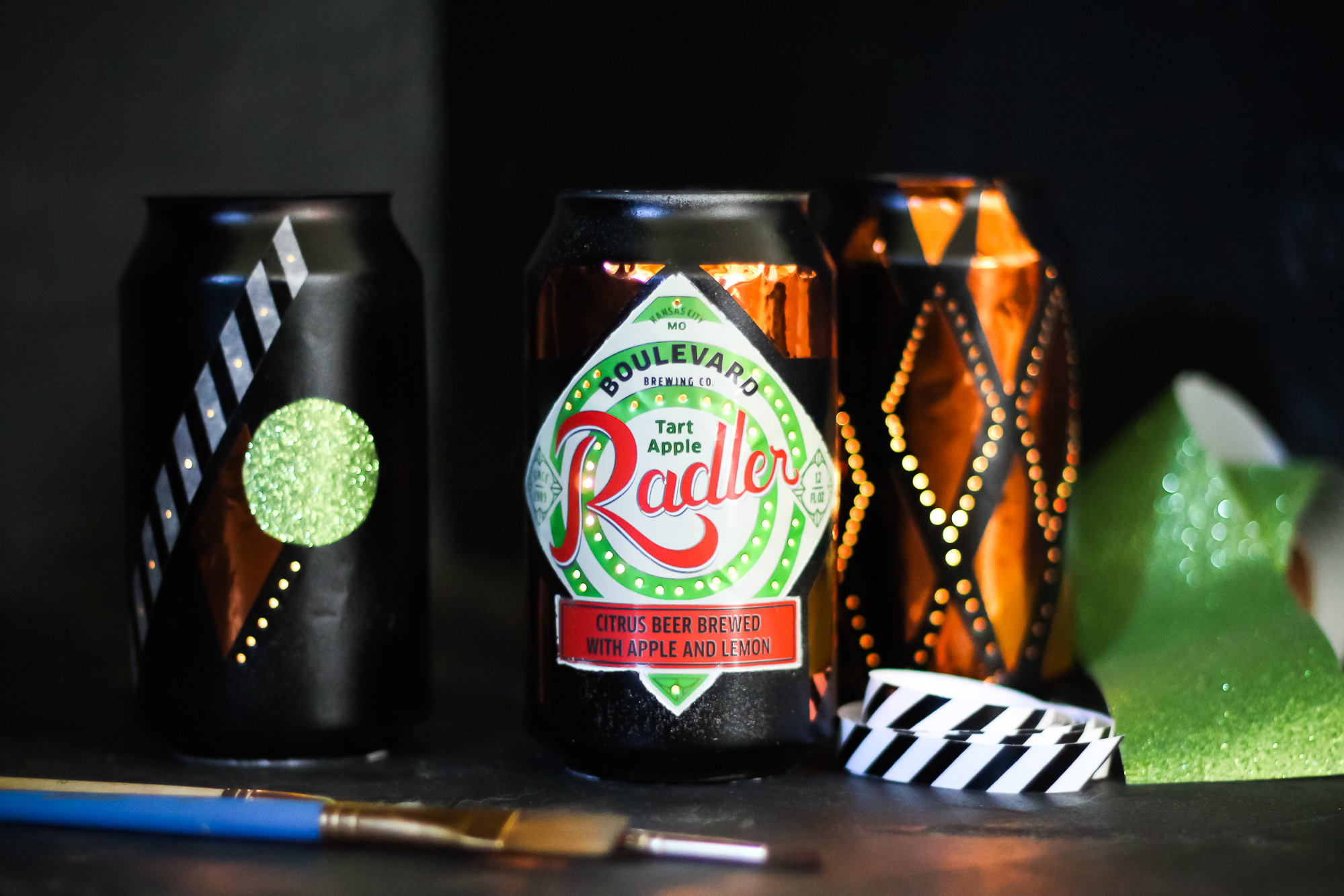 A can of Boulevard Tart Apple Radler is flanked by two decorated black and orange cans with geometric patterns. There’s a paintbrush and green glitter in the foreground, suggesting a DIY craft setup.