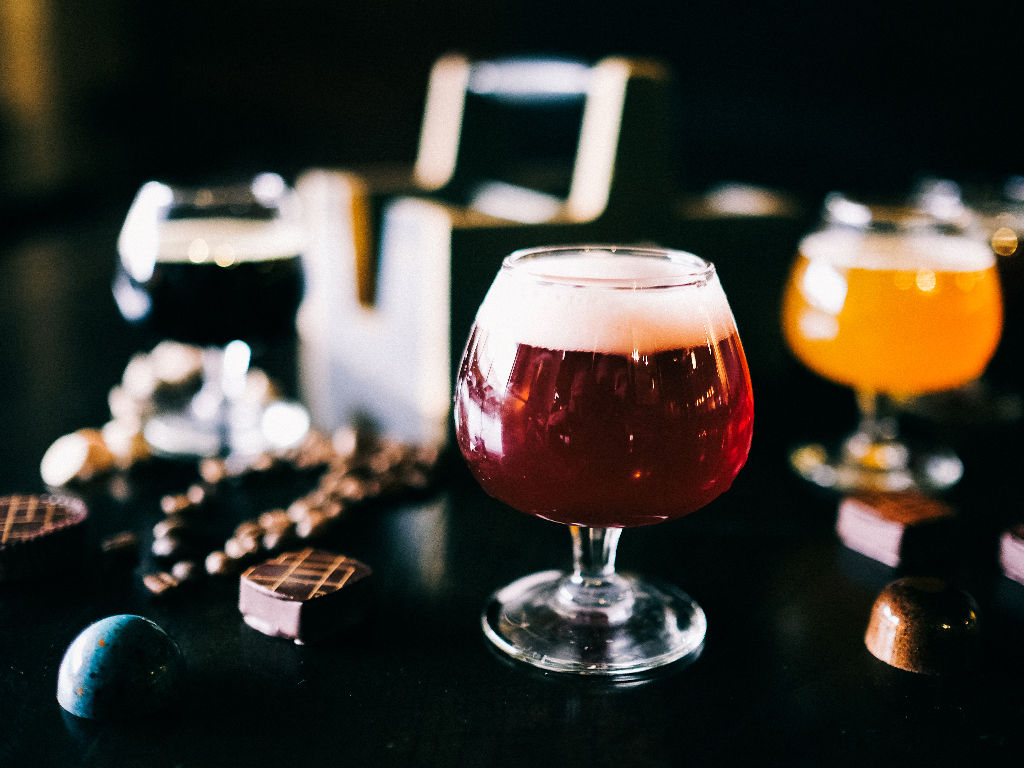 A close-up of a glass of red beer with foam on a dark table, surrounded by chocolates and additional glasses of beer in the background. The scene is blurred and dimly lit, creating a cozy, intimate atmosphere.