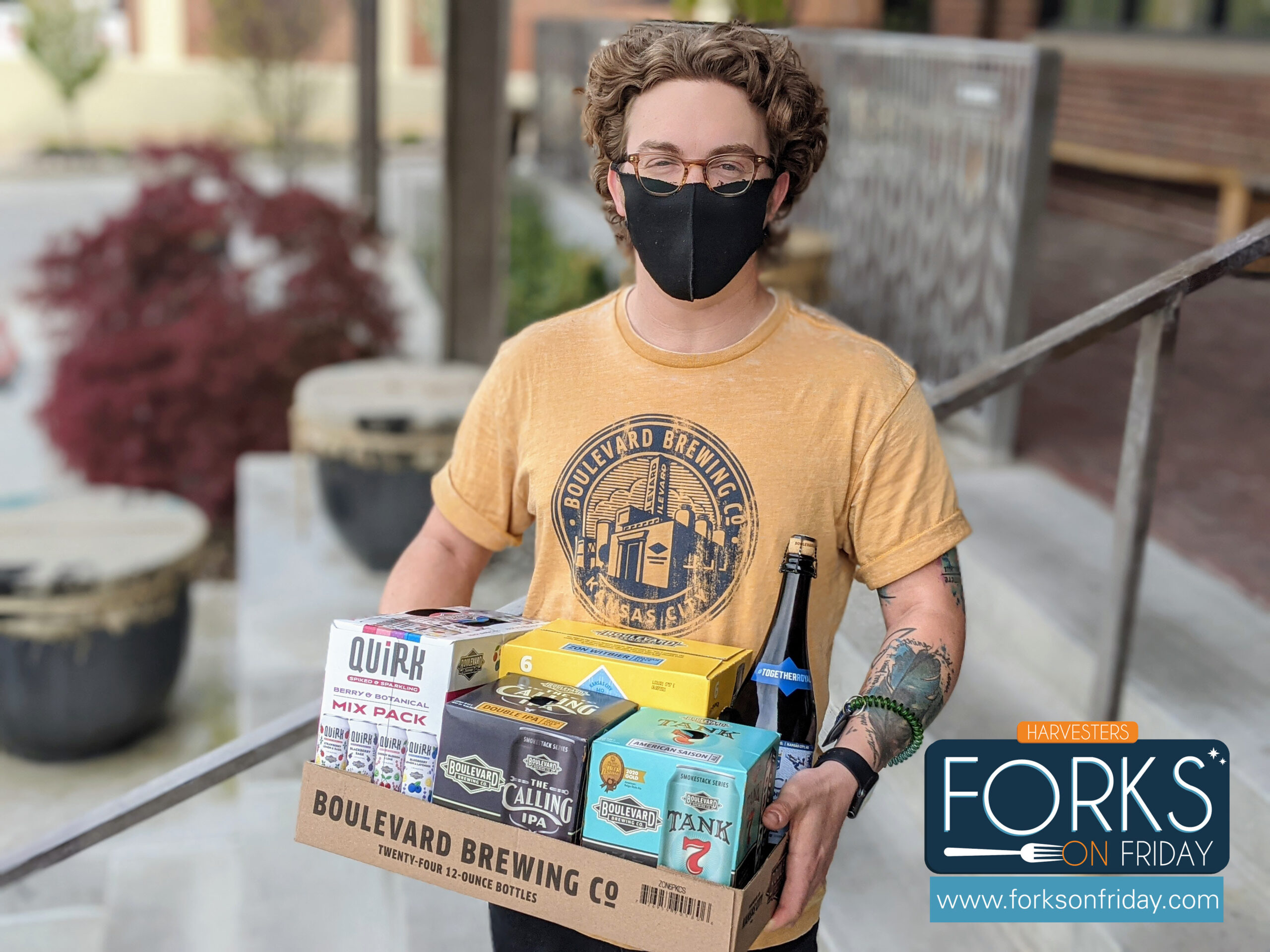A person wearing a black mask, glasses, and a yellow Boulevard Brewing Co. shirt is carrying a box of assorted beers. They are standing on an outdoor staircase. The box includes various beer packs, and a "Forks on Friday" sign is in the bottom right corner.