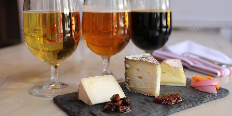 A slate board with assorted cheeses and chutney in the foreground. Three glasses of different colored beers are in the background. A white napkin and cutlery are placed to the side on a wooden table.