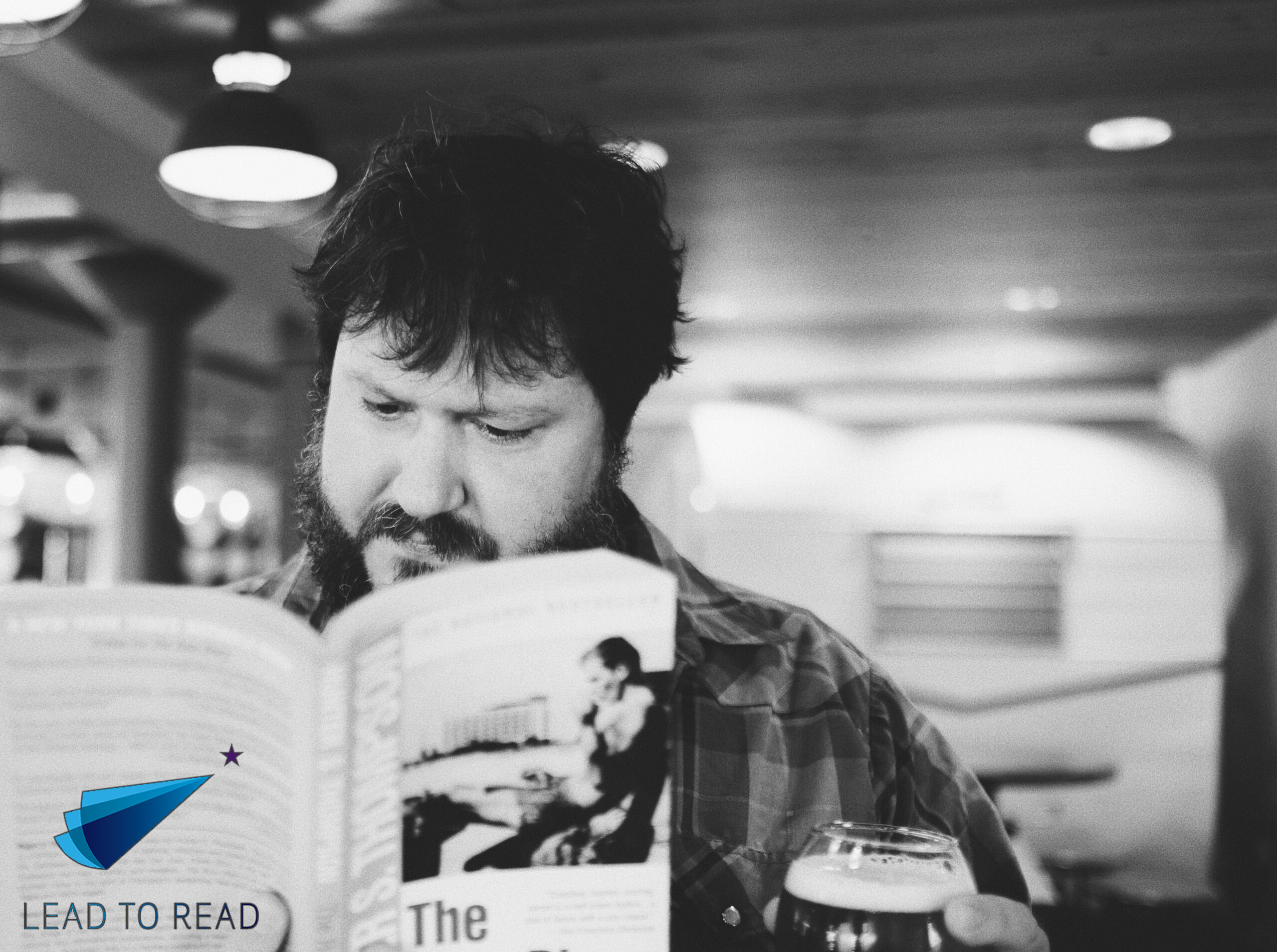A person with a beard is reading a book and holding a glass. The space appears to be a cozy, indoor setting with ambient lighting. A logo with the text "Lead to Read" and a small paper airplane is visible in the corner of the image.
