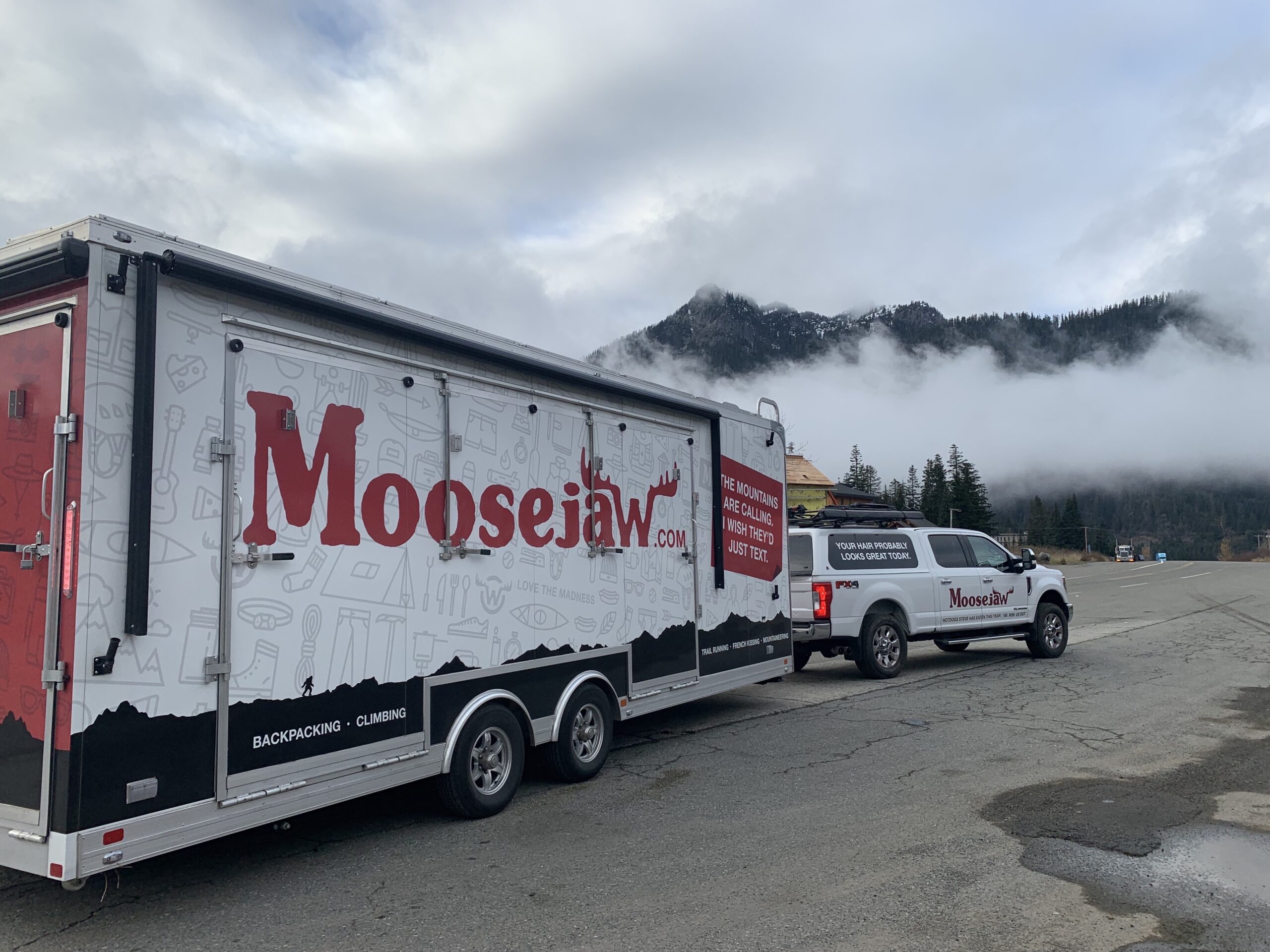 A Moosejaw truck and trailer parked on a cloudy mountain road. The trailer displays the Moosejaw logo and graphics. Pine trees and misty mountains are visible in the background.