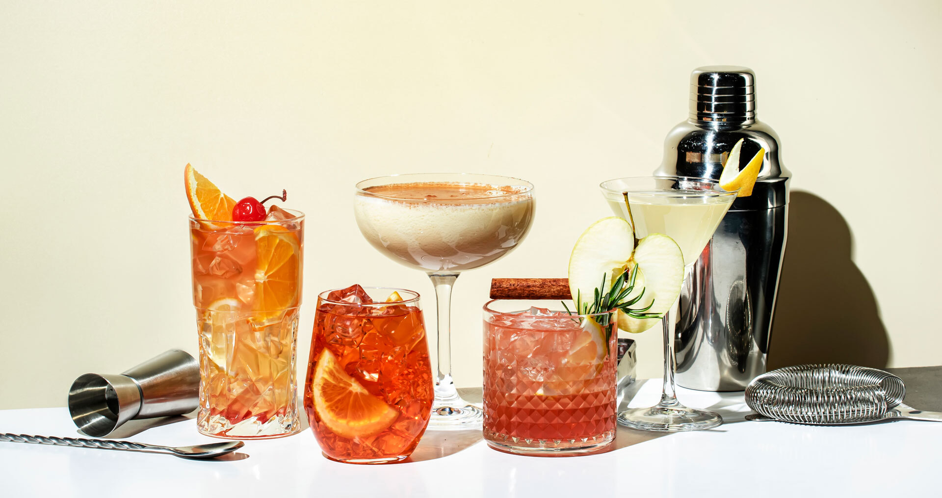 A collection of six different cocktails in various glasses garnished with fruits and herbs, placed on a white surface against a beige background. Accompanied by bar tools, including a shaker, a strainer, a jigger, and a stirring spoon.