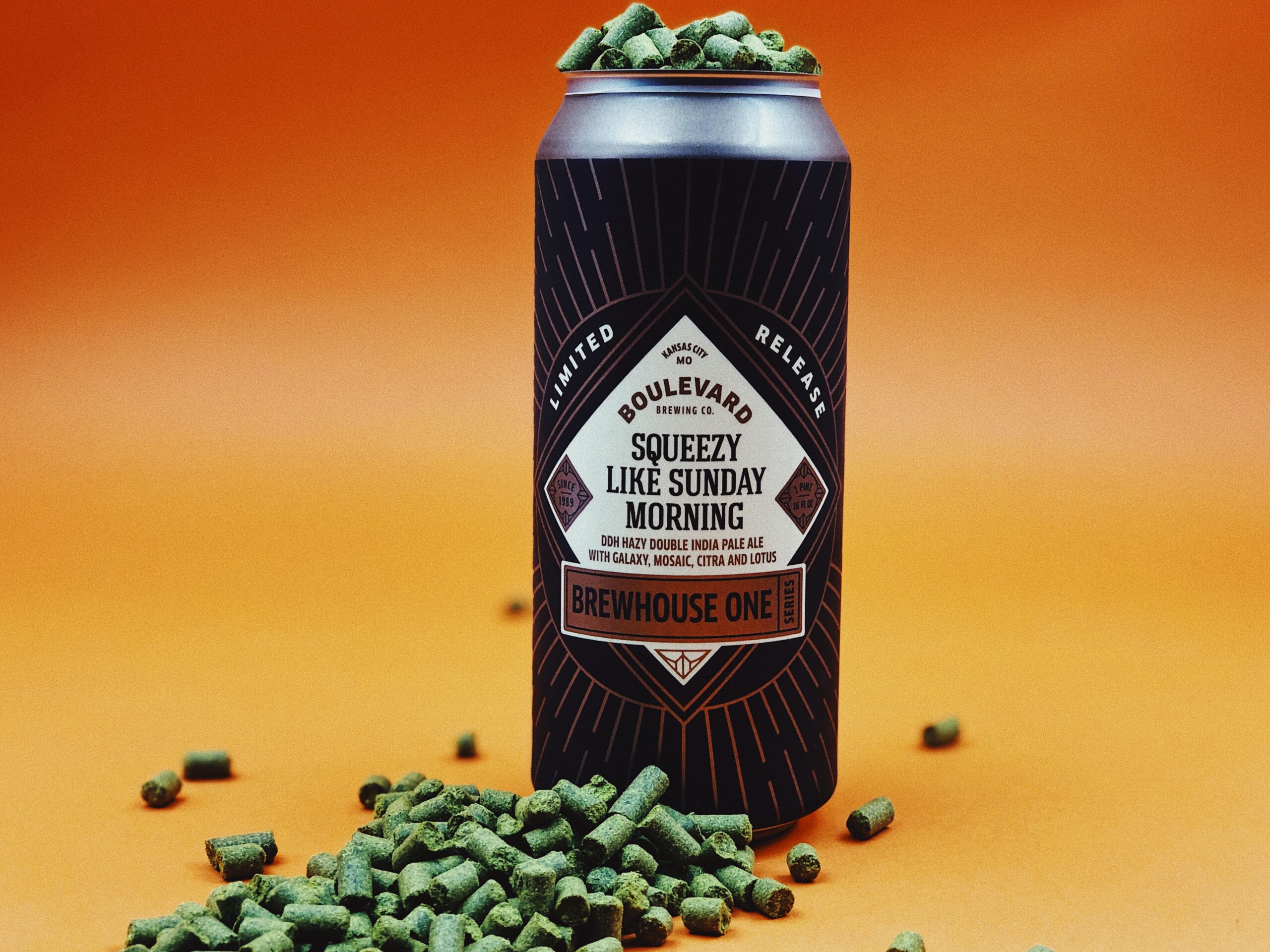 A can of Boulevard Brewing Co.'s "Squeezy Like Sunday Morning" hazy double IPA surrounded by green hop pellets on an orange background. The design features a modern label with focus on the product name and brewery logo.