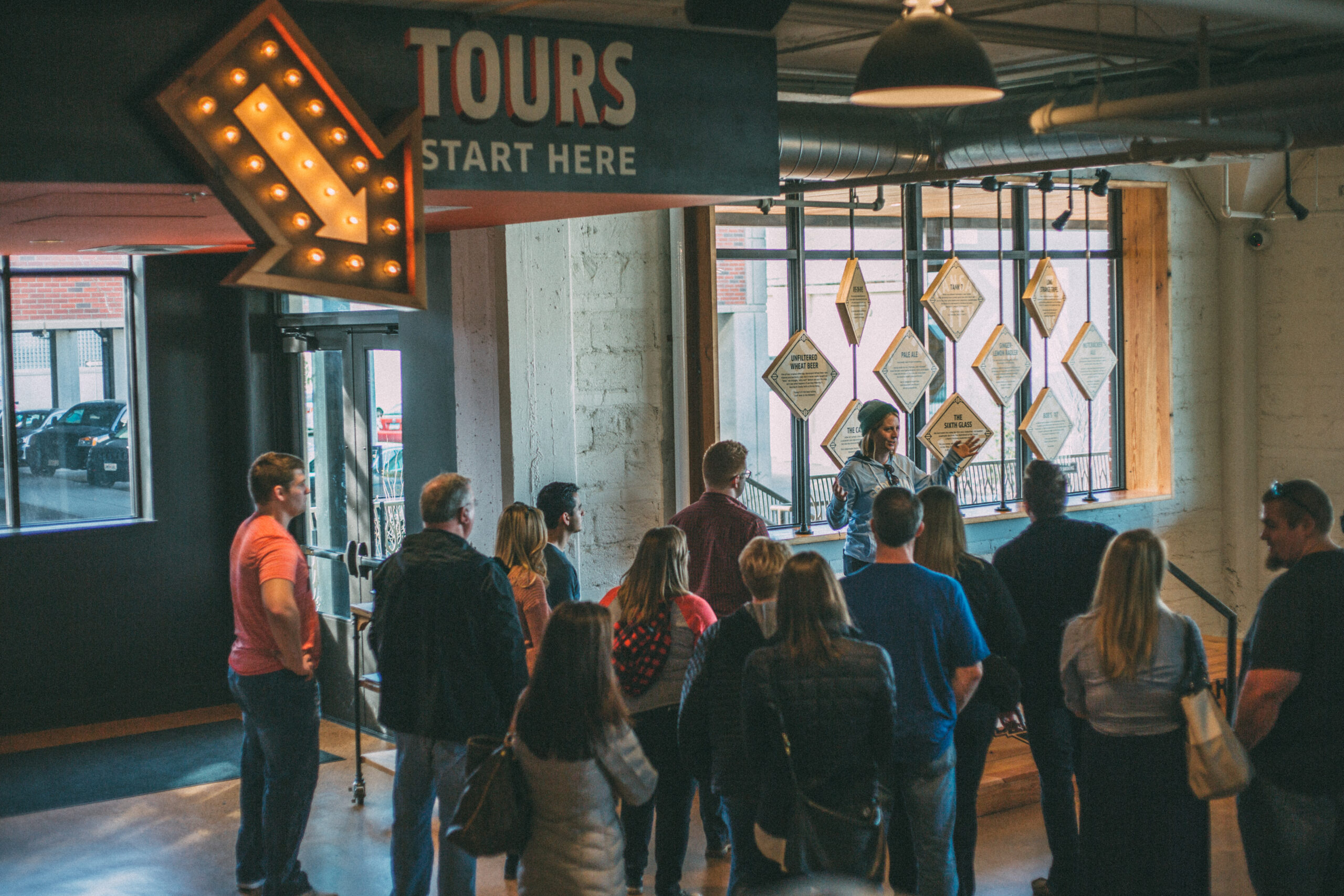 A group of people gather around a tour guide in an indoor setting. A sign above them reads "TOURS START HERE" with an arrow pointing towards the guide. The area has industrial decor with large windows and warm lighting.