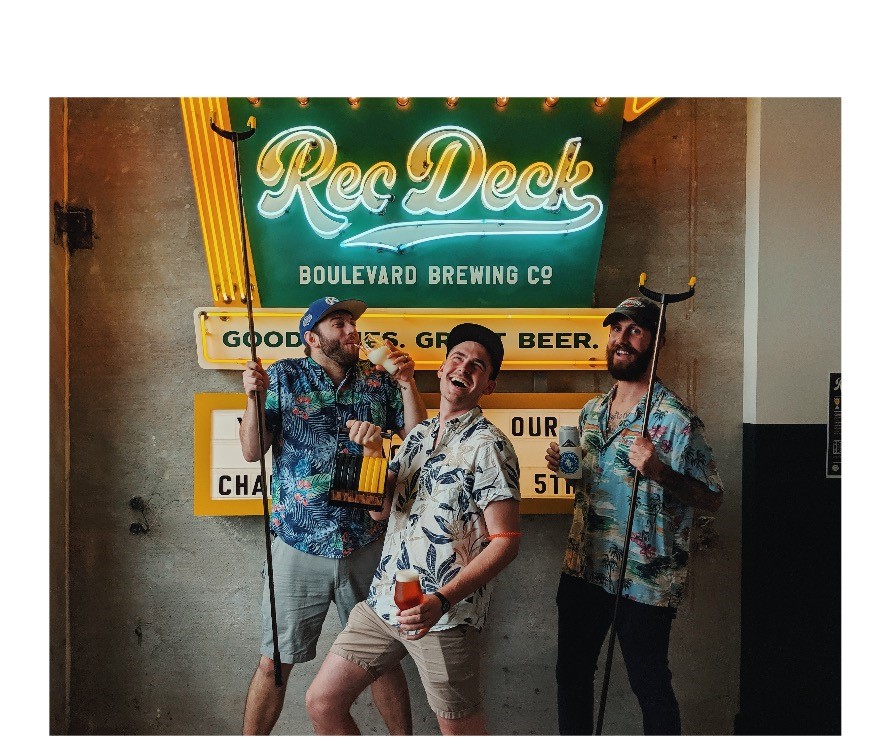 Three people in colorful shirts pose with shuffleboard equipment in front of a neon sign at Boulevard Brewing Co. One holds a drink and smiles broadly, while the others stand on either side, looking at the camera.