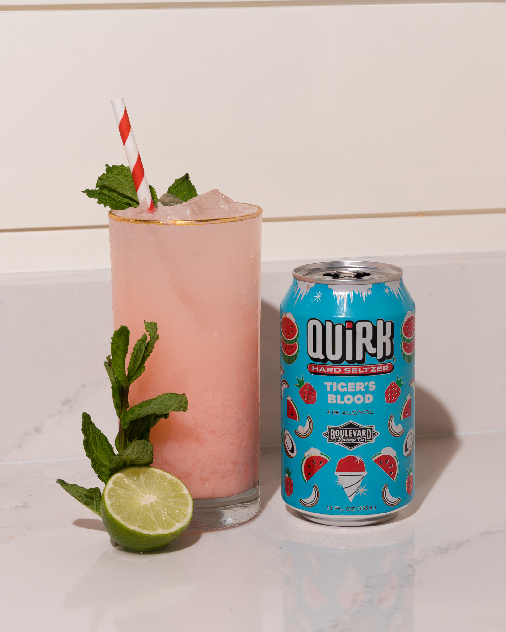 A pink drink with a red-and-white striped straw and mint garnish sits beside a can of Quirk Hard Seltzer (Tiger’s Blood flavor) and a cut lime on a white countertop.