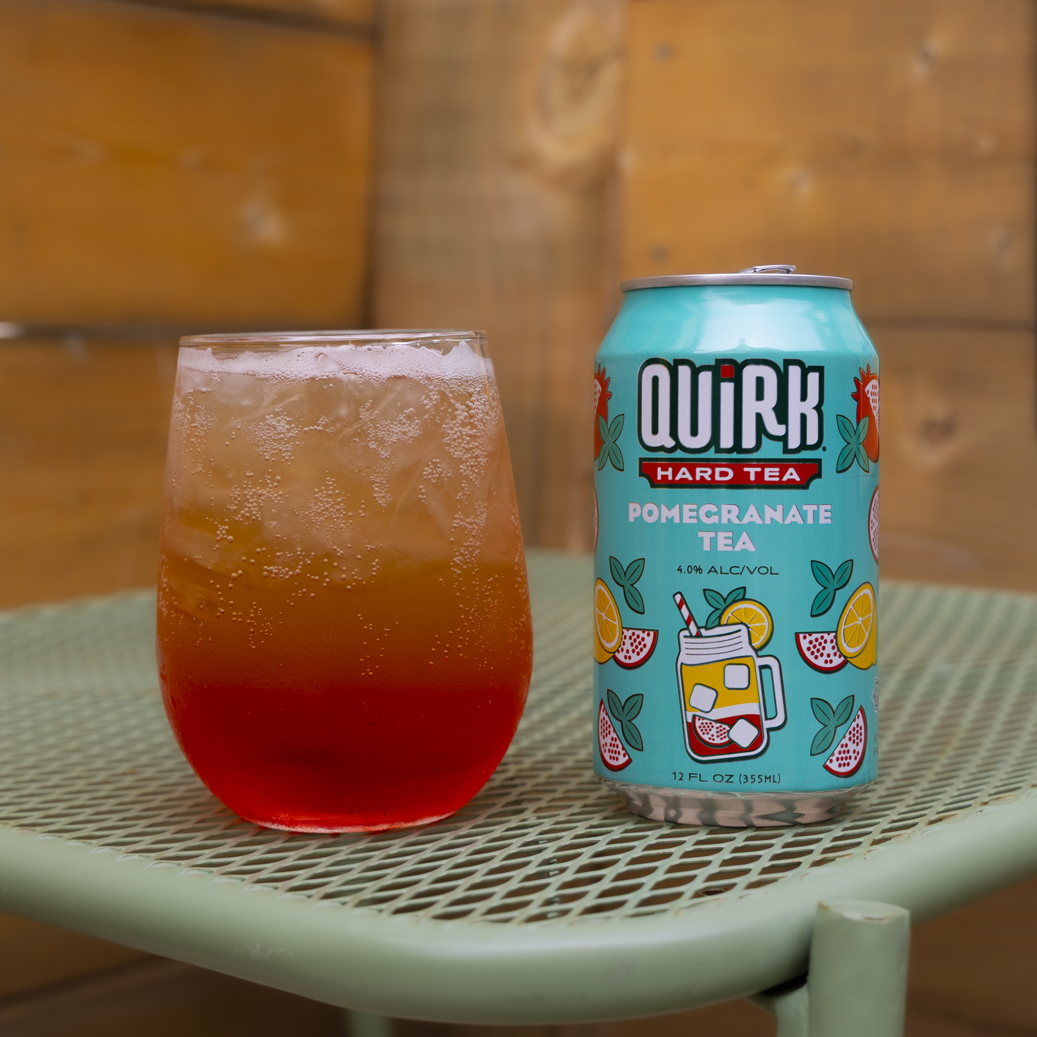 A can of Quirk Hard Tea Pomegranate Tea sits on a green metal table next to a glass filled with ice and a red beverage. The background features a wooden wall.