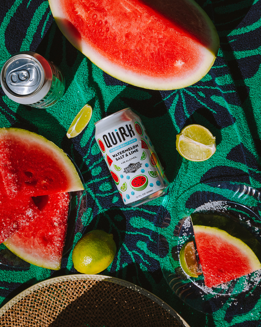 A can of Quirk Watermelon Salt & Lime seltzer is surrounded by watermelon slices, lime wedges, and a whole lime on a green patterned surface, with another can partially visible in the corner.