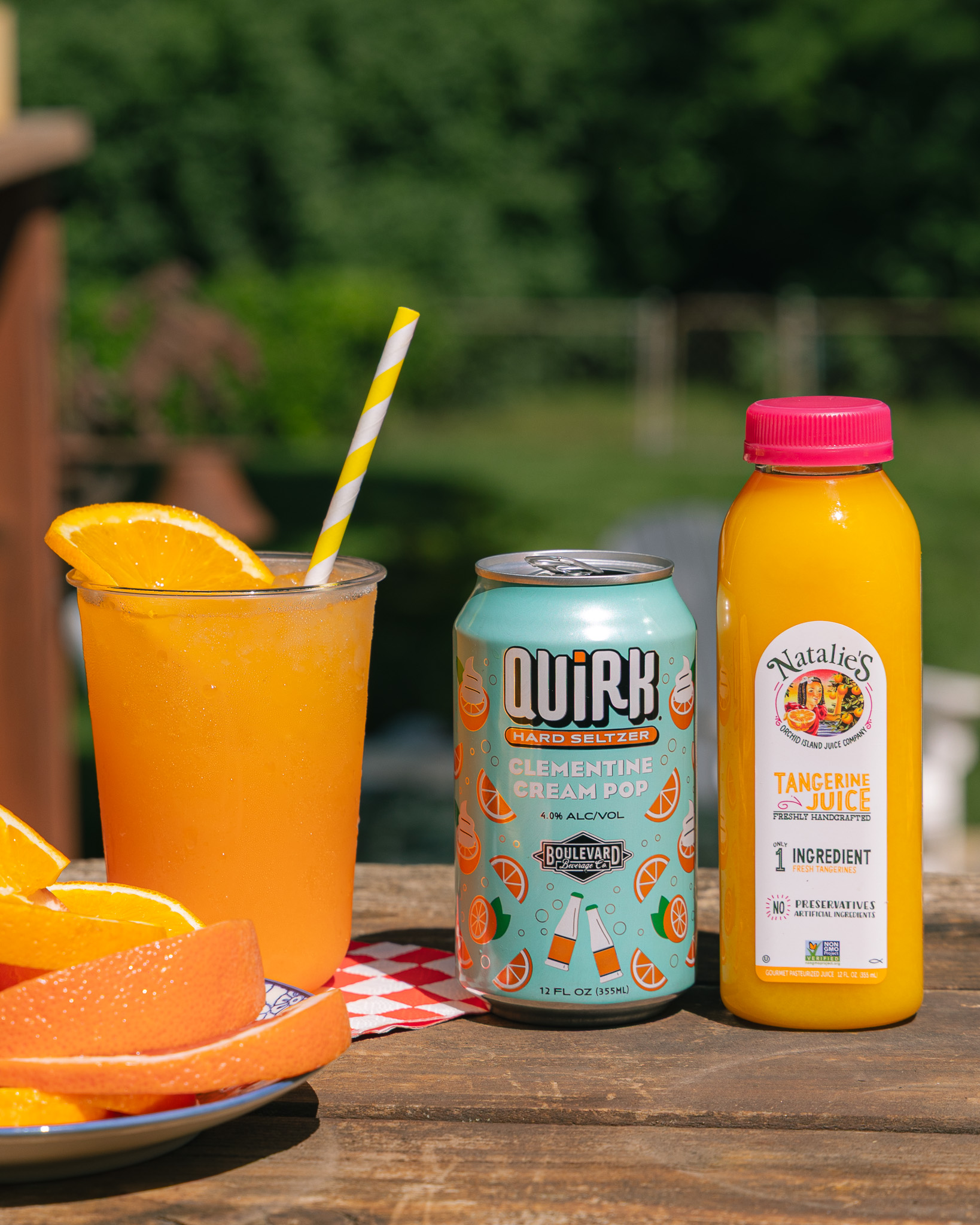 A glass of Tangerine Spritz with a striped straw and orange slice sits on a wooden table next to a can of Quirk Clementine Cream Pop hard seltzer, a bottle of Natalie’s tangerine juice, and a plate of orange slices.
