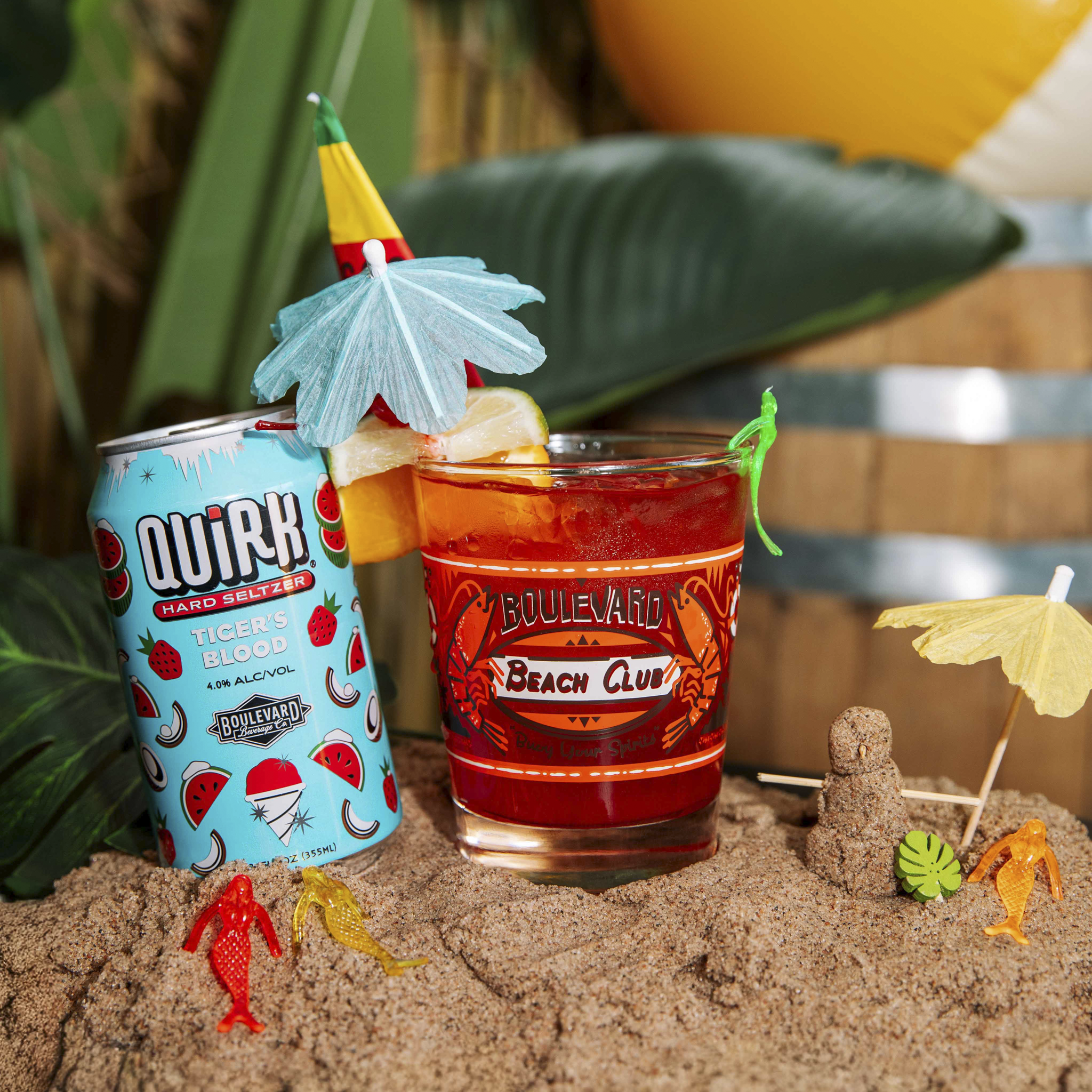 A can of Quirk hard seltzer and a glass with an orange drink, ice, a blue umbrella, lemon wedge, and plastic animals are set on sand with tropical leaves, a sandcastle, and beach decorations in the background.