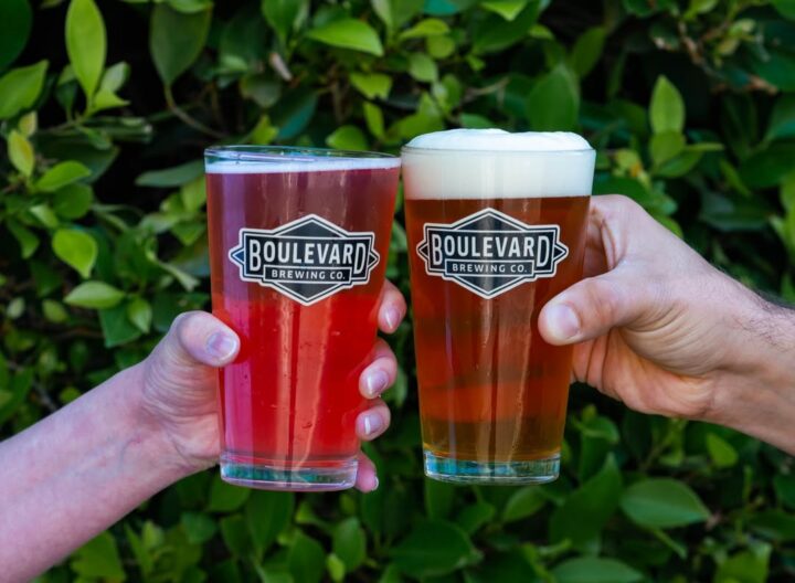 Two hands hold up Boulevard Brewing Co. pint glasses filled with different beers—one red, one golden with foam—against a leafy green background.