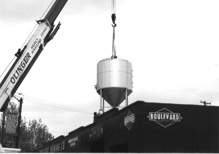 A crane lifts a large, cylindrical tank onto the rooftop of a brick building. Signs on the building read "Boulevard" and "Olinger," indicating the involved businesses. The sky is overcast, and electrical lines are visible in the background, adding to the industrial atmosphere about the scene.