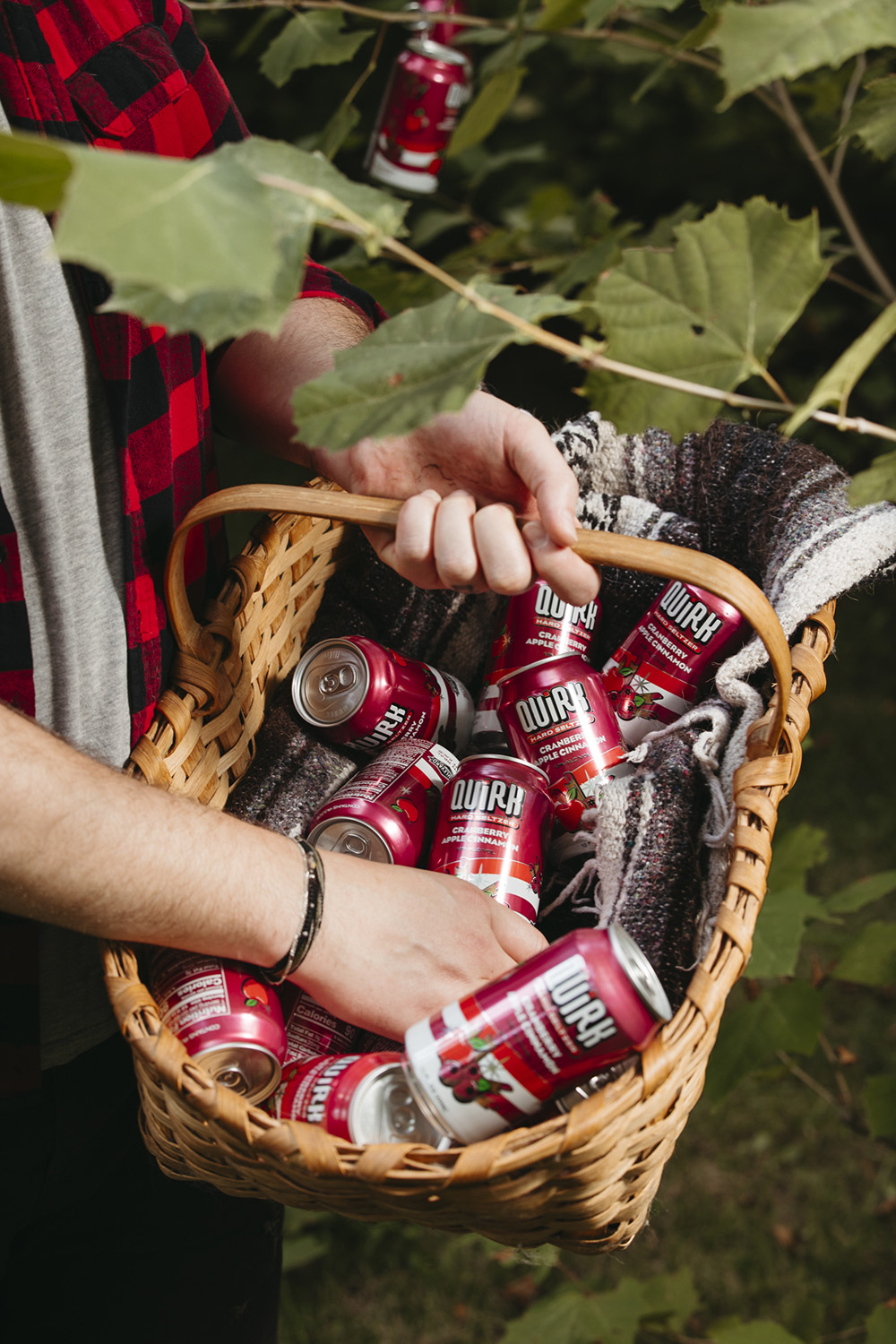 A person wearing a plaid shirt holds a wicker basket filled with red cans labeled "Qurk." The basket rests on their arm amidst green foliage, and a gray and white blanket lines the inside. One of the cans is also hanging on a nearby branch.