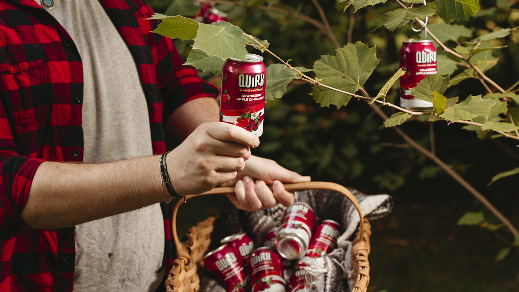 A person in a red and black checkered shirt is holding a can of Quirk Hard Seltzer next to some green branches. Some of the cans are hanging from the branches, while others are in a basket held by the person. The environment appears to be an outdoor, leafy setting.