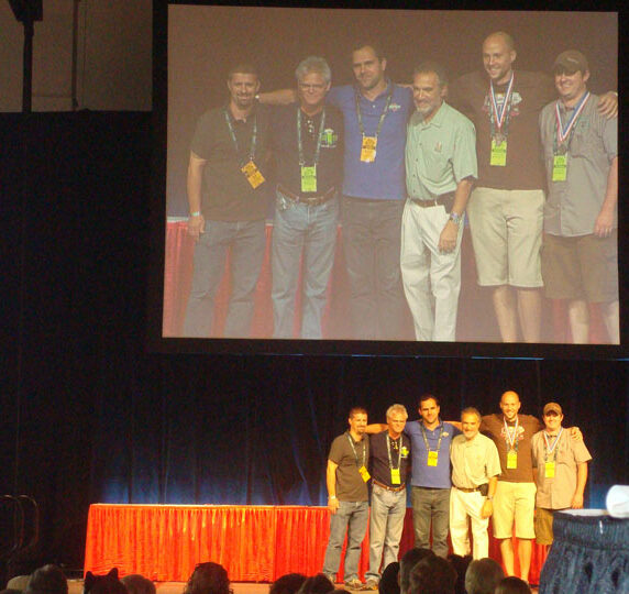 A group of seven men pose together on a stage, with their image projected on a large screen above them. They are standing in front of a red curtain. Audience members are visible in the foreground.