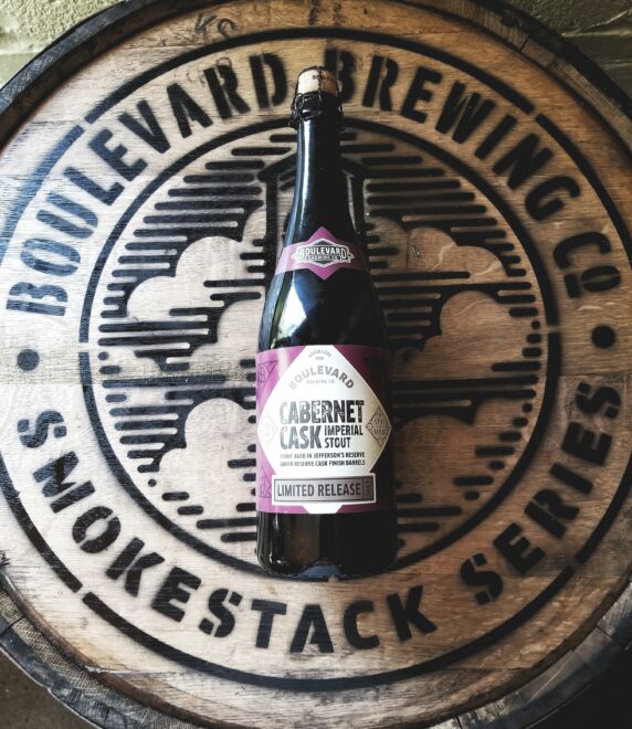 A bottle of Boulevard Brewing Co.'s Cabernet Cask Imperial Stout sits on a wooden barrel with the brewery's logo, featuring "Smokestack Series" and "Limited Release" branding.