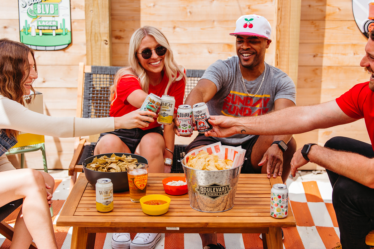 Four friends are enjoying a casual outdoor gathering. They are seated around a small table with snacks and drinks, toasting with cans of beer. The setting is relaxed, with a wooden fence in the background and chairs for seating.