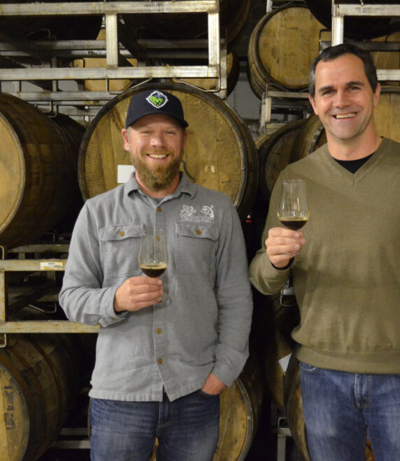 Two men smiling and holding wine glasses stand in front of stacked wooden barrels. One wears a cap and a grey shirt, the other a brown sweater. They appear relaxed in a rustic setting.