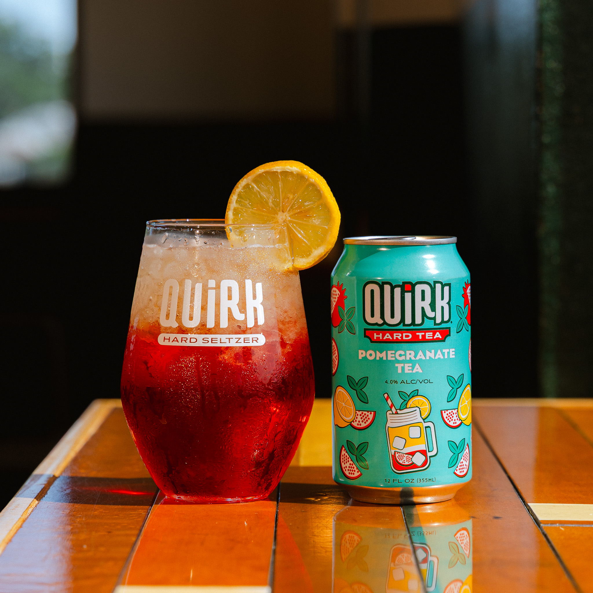 A glass of red Quirk Hard Seltzer with ice and a lemon slice sits next to a colorful can labeled “Quirk Hard Tea Pomegranate Tea” on a wooden table.
