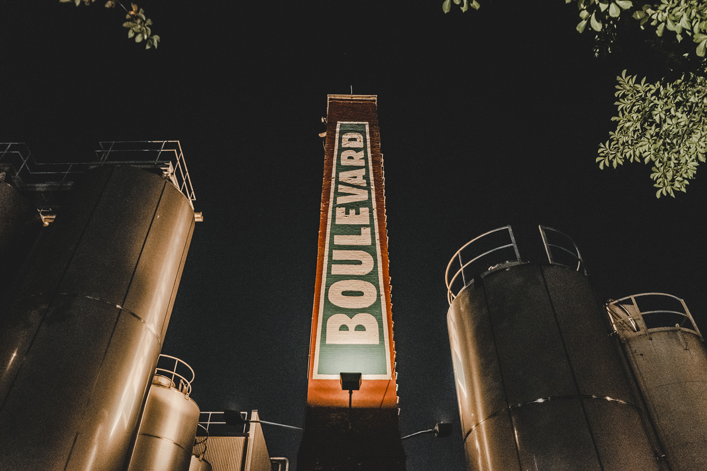 A tall, illuminated sign displaying "Boulevard" is flanked by large, metal silos. The scene is set against a dark night sky, with tree branches partially visible on the upper right corner.