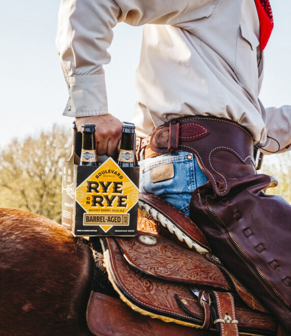 A person wearing a beige shirt and blue jeans, seated on a horse, holds a six-pack of Rye beer in their right hand. The rider is equipped with traditional cowboy gear, including chaps and a belt, with trees and bright sky in the background.