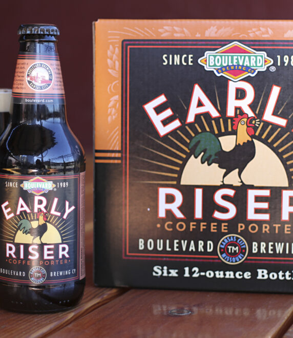 A bottle and six-pack of Boulevard Brewing Co.'s Early Riser Coffee Porter are displayed on a wooden table. The bottle is next to a filled glass showcasing the dark beer. The label features a rooster and vintage-style design.