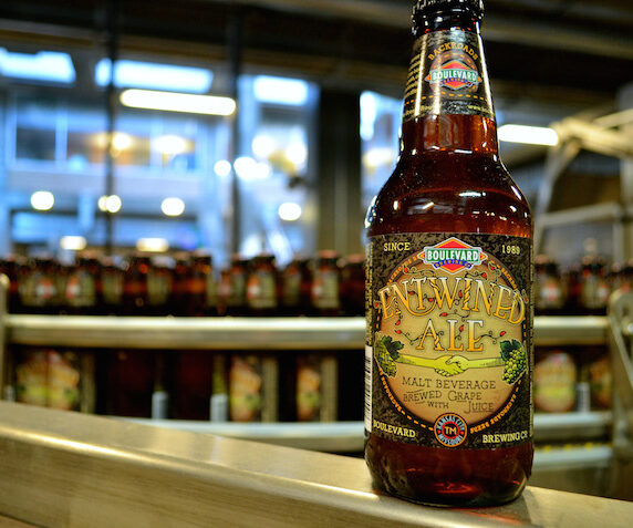 A bottle of Boulevard's "Funkier Ale" with a colorful label sits in the foreground on a conveyor belt in a brewery. The background shows rows of similar bottles in a well-lit industrial setting.