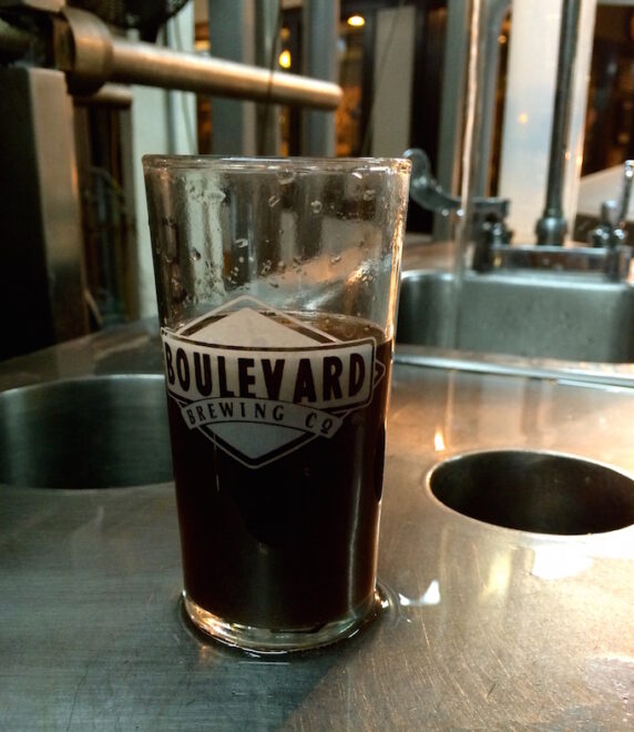 A half-full glass of dark beer with the logo "Boulevard Brewing Co" sits on a metal surface in a brewery setting, featuring stainless steel equipment in the background.