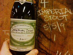 A hand holding a bottle of Imperial Stout with a detailed label. In the background, "Imperial Stout 5/9/9" is written in chalk on a wooden surface.