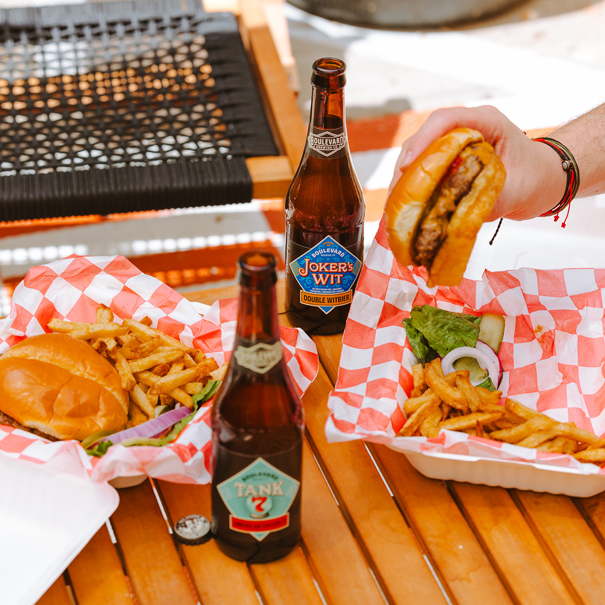 Two plates of burgers and fries are on a wooden table covered with red and white checkered paper. Two beer bottles, labeled "Joker's Wit" and "Tank 7," are on the table. A hand is holding up one of the burgers above the table. A cap is next to a bottle.