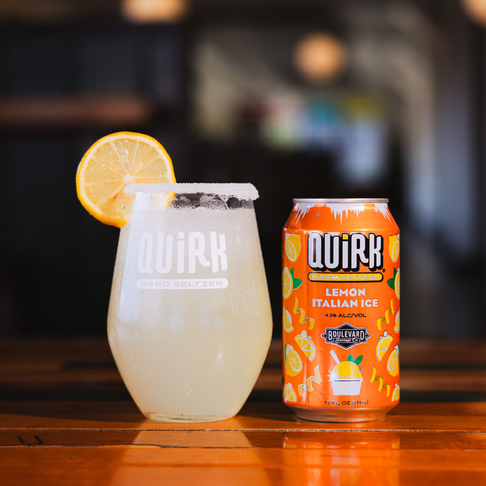 A glass of Quirk hard seltzer with a sugar rim and lemon slice is next to a can of Quirk Lemon Italian Ice hard seltzer on a wooden table.