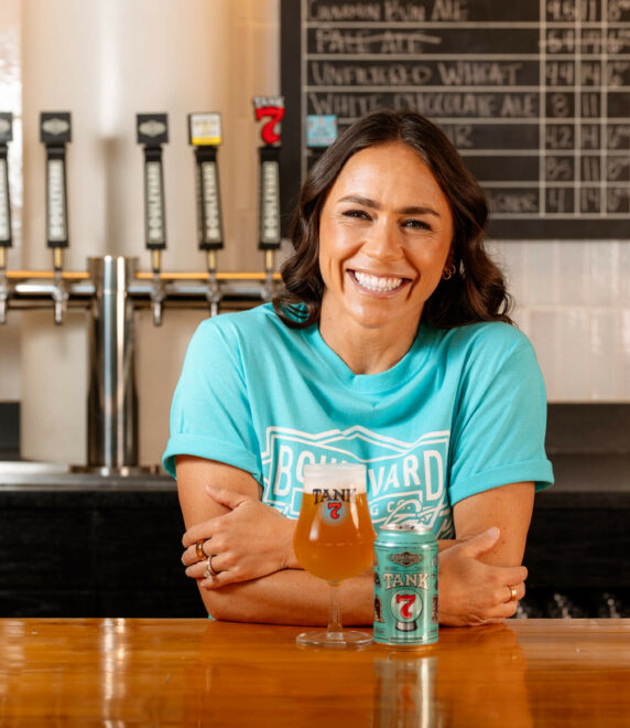 A person with long dark hair and a big smile wears a turquoise T-shirt and sits at a wooden bar. A beer glass and a branded beer can are on the bar in front of them, and beer taps and a chalkboard with drink options are visible in the background.