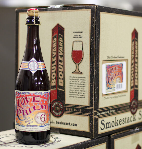 A bottle of "Love Child No. 6" beer stands in front of a box that features the same label design. The label is colorful with red and yellow accents. The box is part of the Smokestack Series from Boulevard Brewing Co.