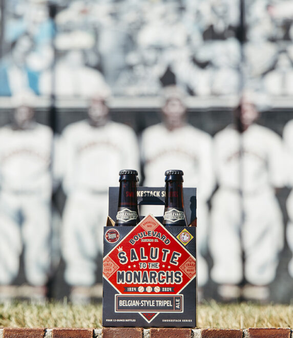 A pack of Boulevard Brewing Company "Salute to the Monarchs" Belgian-style tripel beer is placed on a ledge with a blurred historic baseball team photo in the background. The beer pack features a red, diamond-shaped design and two bottles are visible.