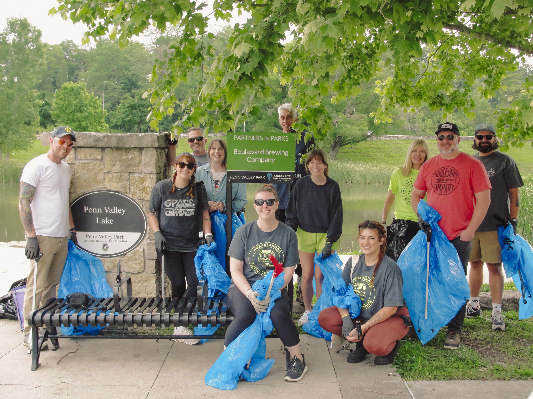 A group of people holding blue trash bags pose in front of a sign for Penn Valley Lake, next to a bench and trees, during a park cleanup event.