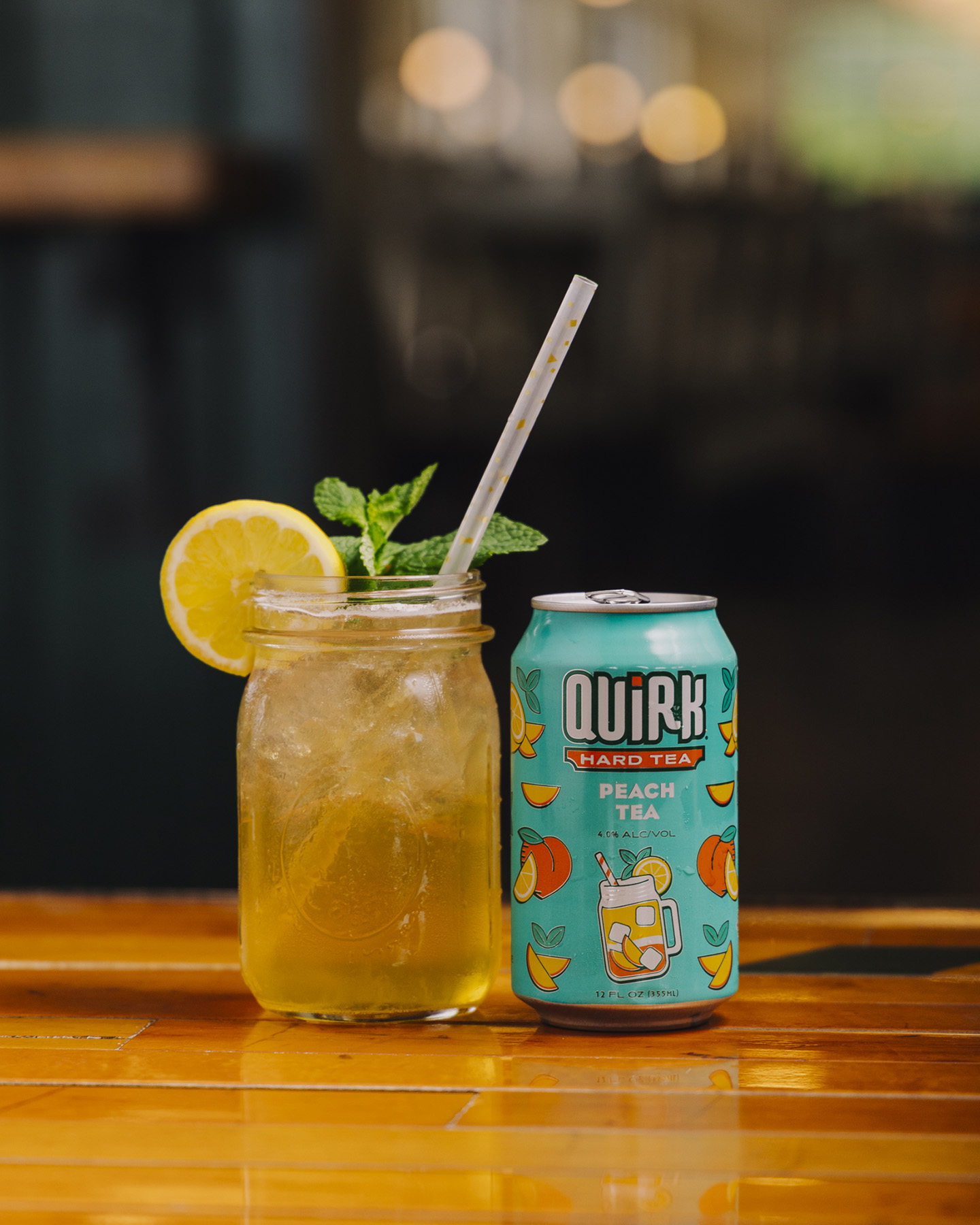 A can of Quirk Peach Tea Hard Tea sits next to a mason jar filled with iced tea, a lemon slice, mint sprig, and a straw, on a reflective wooden surface with a blurry background.