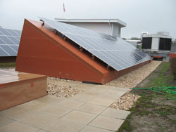 Rooftop view with solar panels mounted on a slanted, rust-colored metal structure. The panels are oriented to capture sunlight. The roof is covered with pebbles and tiles, with a green hose visible to the right.