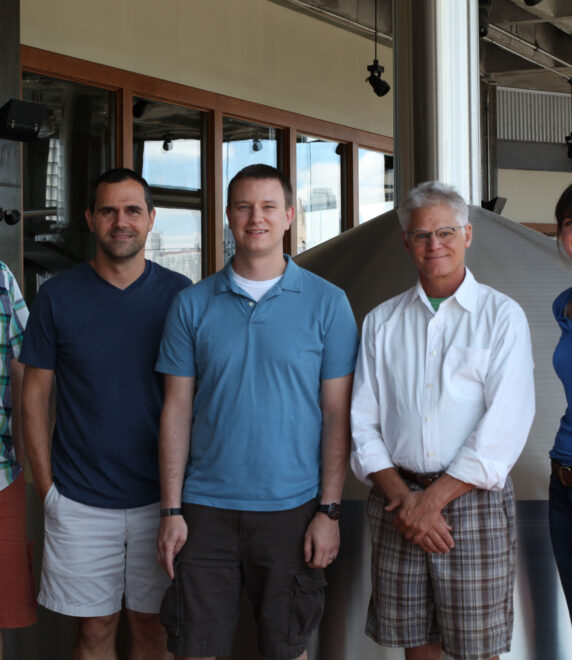 Five people standing together indoors, posing for a photo. They are casually dressed, and behind them is industrial equipment, possibly in a brewery or similar setting. The group includes four men and one woman, all smiling.