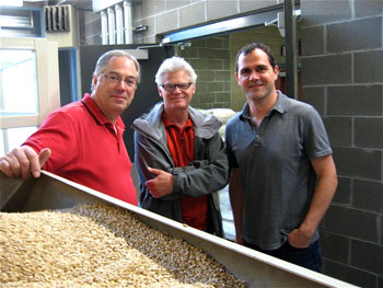 Three men stand together in a room with gray brick walls. In the foreground, there's a large tray filled with grains. The men are smiling, with one wearing a red shirt, another in a jacket, and the third in a gray shirt.