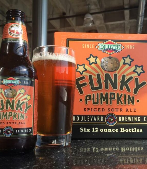 A bottle and a glass of Funky Pumpkin Spiced Sour Ale are placed in front of a six-pack box on a reflective surface, with large windows in the background. The label is orange with bold text and a pumpkin illustration.