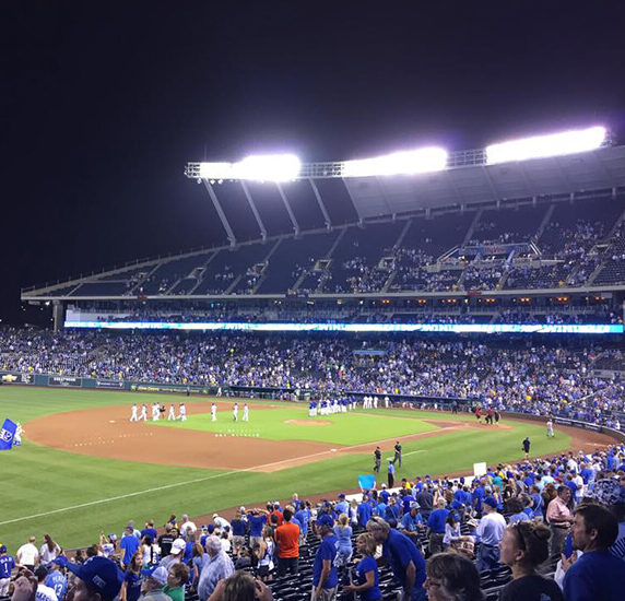 A large crowd gathers in a brightly lit baseball stadium at night. Players in white uniforms are seen on the field, while spectators, many in blue, watch from the stands. The atmosphere is lively, and the stadium is bustling with activity.