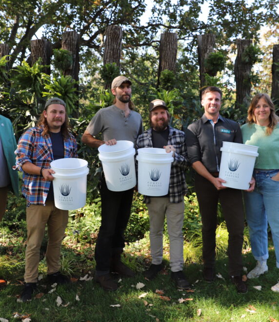 Six people stand outdoors smiling at a camera, surrounded by greenery. Four of them hold white buckets with a black logo. The background features trees and lush foliage.