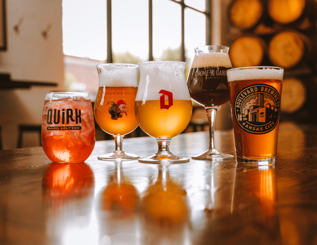 A variety of five craft beer glasses on a wooden table in a brewery setting. Each glass contains a different type of beer, ranging from light to dark, with distinct logos. Large barrels are visible in the background next to a window.